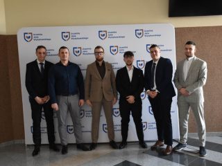 Absolwenci w garniturach stoją w rzędzie przed ścianką UJW.