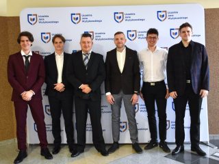 Grupa absolwentów kierunków inżynierskich stoi w rzędzie przy ściance UJW.