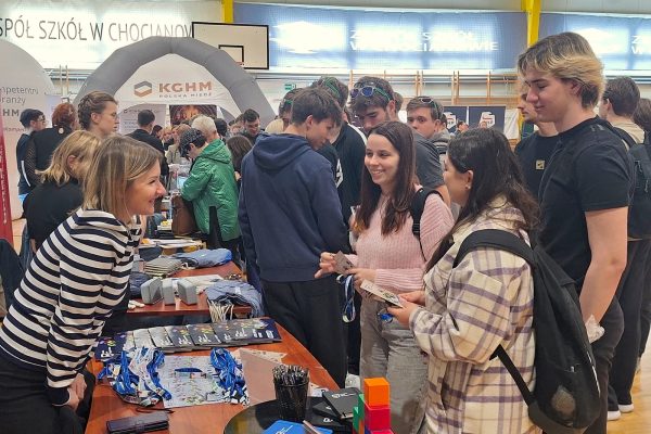 Uczniowie rozmawiają z przedstawicielką uczelni przy stole z ulotkami i gadżetami.