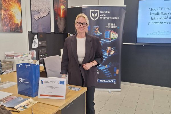 Przedstawicielka UJW przy stoliku z materiałami i ekranie prezentacji w szkole.