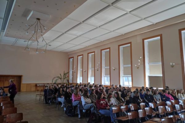 Widok szkolnej auli z uczniami i prowadzącą spotkanie UJW.