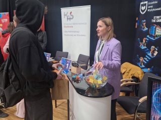 Przedstawicielka UJW w fioletowej marynarce rozmawia z odwiedzającym przy stoisku.