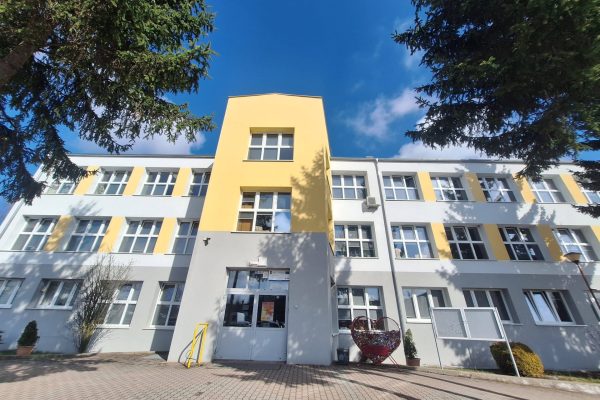 Budynek szkoły z żółto-białą elewacją i drzewami przed wejściem.