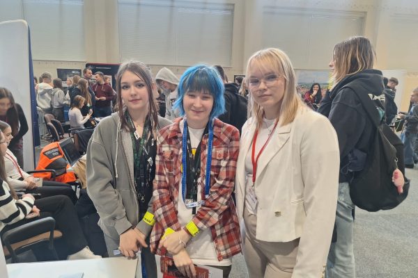 Trzy uczestniczki pozują wśród stoisk na targach edukacyjnych.