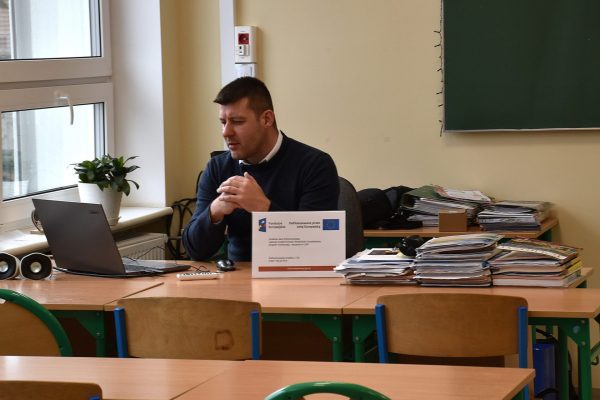 Prowadzący z mikrofonem siedzi przy laptopie w sali lekcyjnej.