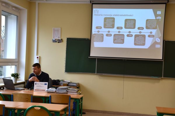 Prowadzący przy laptopie omawia slajd o historii rozwoju elektromobilności.