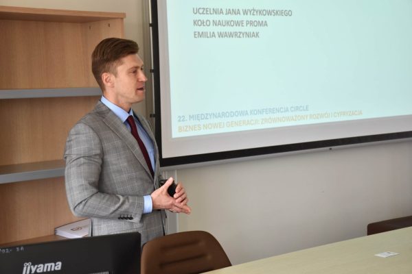 Mężczyzna w szarej marynarce przemawia obok ekranu prezentacji.