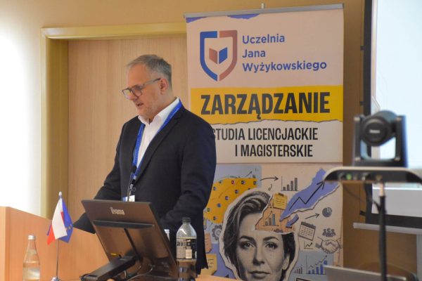 Prelegent w okularach przy laptopie i mównicy na tle roll-upu UJW.