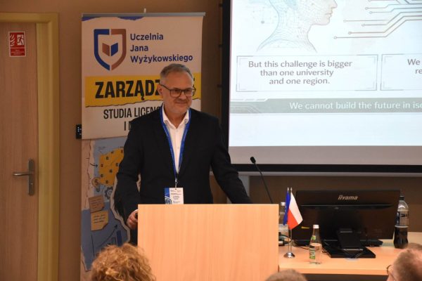 Mężczyzna w ciemnym garniturze przemawia przy mównicy na tle slajdu.