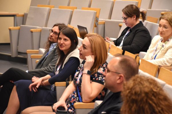 Grupa słuchaczy z identyfikatorami w sali wykładowej podczas konferencji.