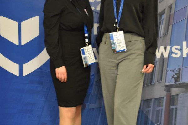 Dwie uczestniczki z identyfikatorami pozują na tle niebieskiej ścianki UJW.