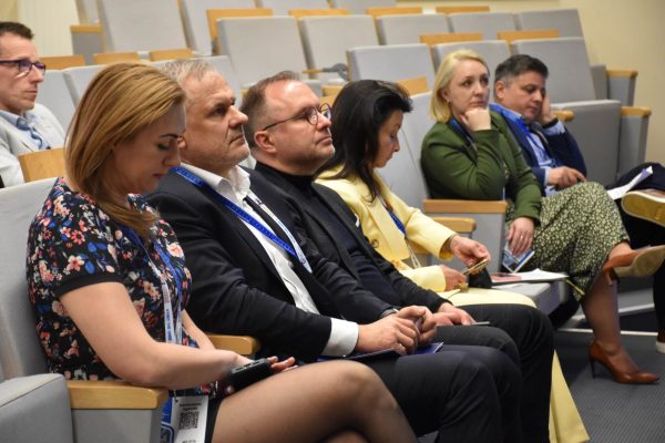 Rząd uczestników konferencji słuchających prezentacji w sali wykładowej.