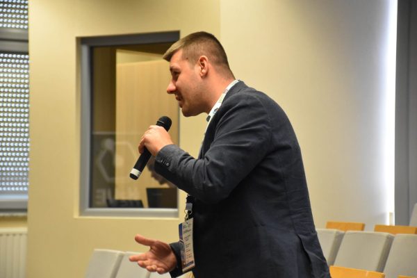 Uczestnik zabiera głos, stojąc z mikrofonem wśród foteli widowni.
