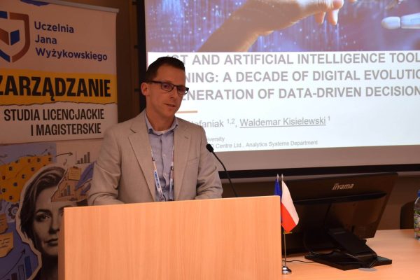 Zbliżenie na prelegenta w jasnej marynarce przy mównicy i ekranie prezentacji.