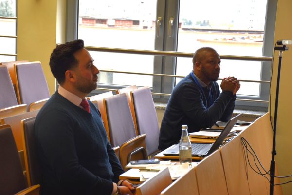 Dwaj uczestnicy konferencji słuchają prezentacji przy otwartych laptopach.