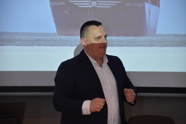 Mężczyzna prowadzi prezentację na tle zdjęcia statku wyświetlonego na ekranie.