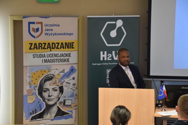 Prelegent w garniturze przy mównicy między planszami UJW i H2VE.