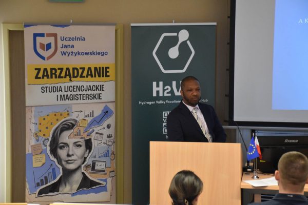 Mężczyzna w garniturze przemawia przy mównicy na tle planszy H2VE.