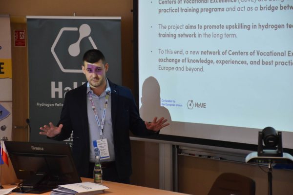 Prelegent gestykuluje przy ekranie prezentacji projektu H2VE.