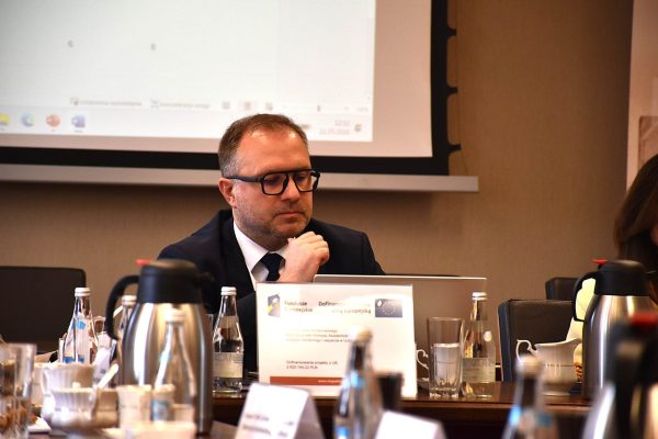 Mężczyzna w okularach słucha dyskusji przy laptopie.