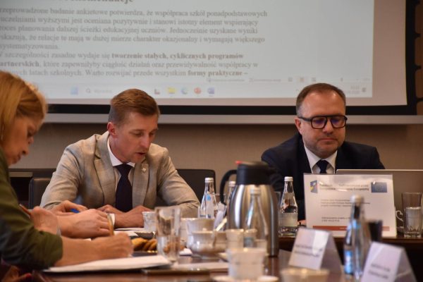 Uczestnicy posiedzenia pracują przy stole na tle dokumentu wyświetlonego na ekranie.