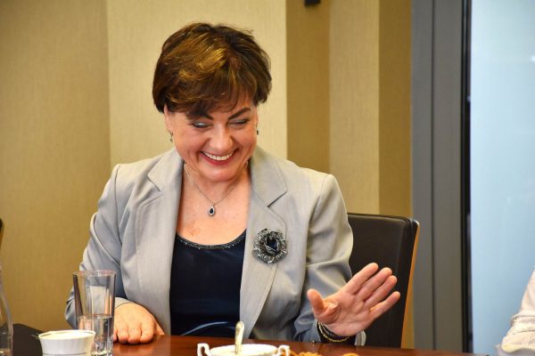 Uśmiechnięta uczestniczka w szarej marynarce gestykuluje przy stole.