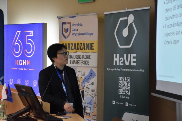 Prelegent w okularach stoi przy komputerze obok roll-upu H2VE.