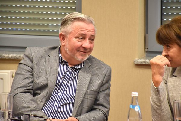 Uśmiechnięty uczestnik w szarej marynarce rozmawia z sąsiadką.