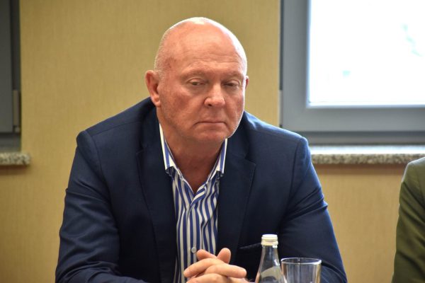 Uczestnik w granatowej marynarce słucha posiedzenia ze splecionymi dłońmi.