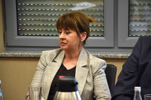 Uczestniczka w szarej marynarce słucha dyskusji przy stole.