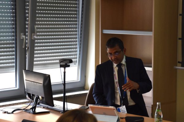 Prelegent spogląda na komputer, stojąc przy mikrofonie.