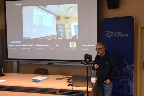 Mężczyzna stoi przy kamerze pod ekranem zdalnego połączenia konferencyjnego.