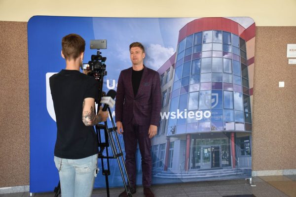 Operator kamery nagrywa wypowiedź mężczyzny w bordowym garniturze.