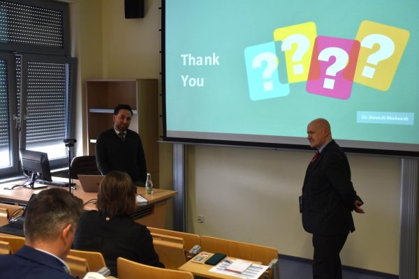 Dwaj uczestnicy rozmawiają przed publicznością pod slajdem z podziękowaniem i znakami zapytania.