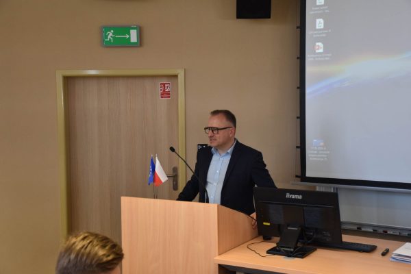 Mężczyzna w okularach otwiera konferencję przy mównicy.