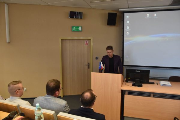 Prelegent w bordowym garniturze występuje przed publicznością.