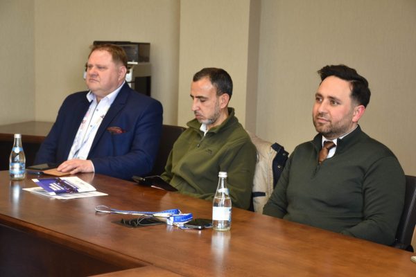 Trzej uczestnicy siedzą przy stole z materiałami konferencyjnymi.