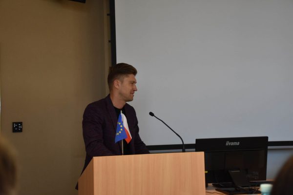 Mężczyzna w bordowym garniturze przemawia przy mównicy.