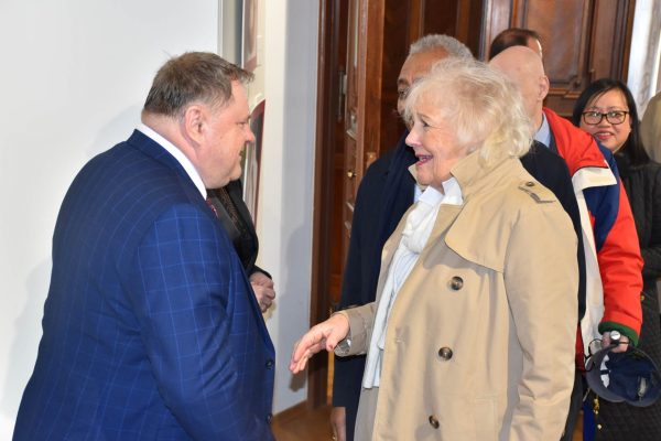 Mężczyzna w garniturze wita uczestniczkę w beżowym płaszczu.