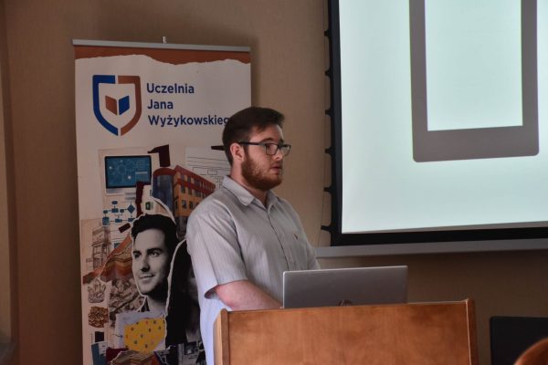Zbliżenie na prelegenta w okularach stojącego przy laptopie.