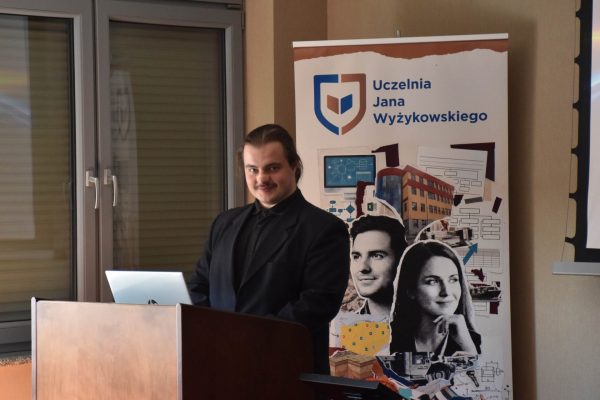 Prelegent w czarnej koszuli stoi przy laptopie na mównicy.