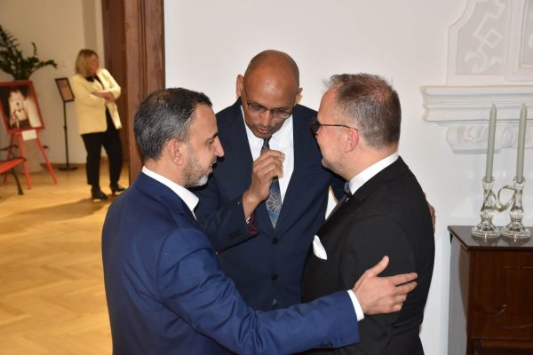 Trzej mężczyźni w garniturach rozmawiają w sali uroczystej kolacji.