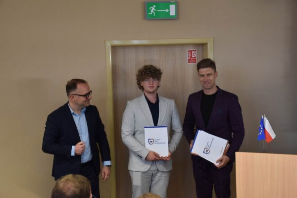 Uczestnik w jasnej marynarce prezentuje dokument obok organizatorów.