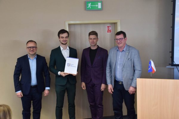 Uczestnik w zielonym garniturze pozuje z dokumentem wraz z organizatorami.