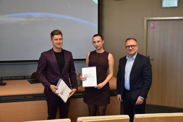 Uczestniczka w bordowej sukience prezentuje dokument przy organizatorach.