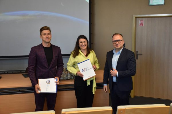 Uczestniczka w zielonej bluzce pozuje z dokumentem obok organizatorów.