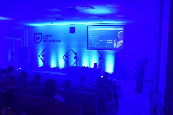 Aula UJW oświetlona na niebiesko, z ekranem konferencji i publicznością.