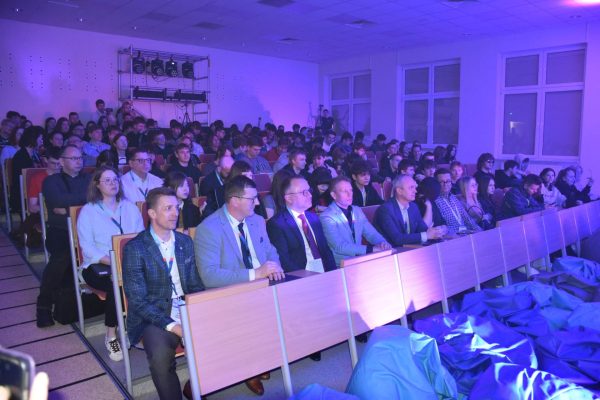 Uczestnicy konferencji siedzą w rzędach w auli oświetlonej na fioletowo.