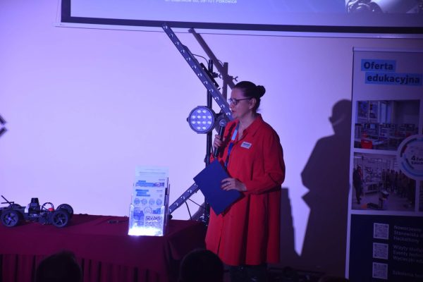 Prowadząca w czerwonej sukience z mikrofonem i teczką na scenie konferencji.