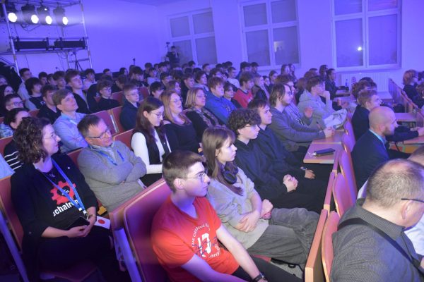 Publiczność słucha wystąpienia, siedząc przy pulpitach w auli.